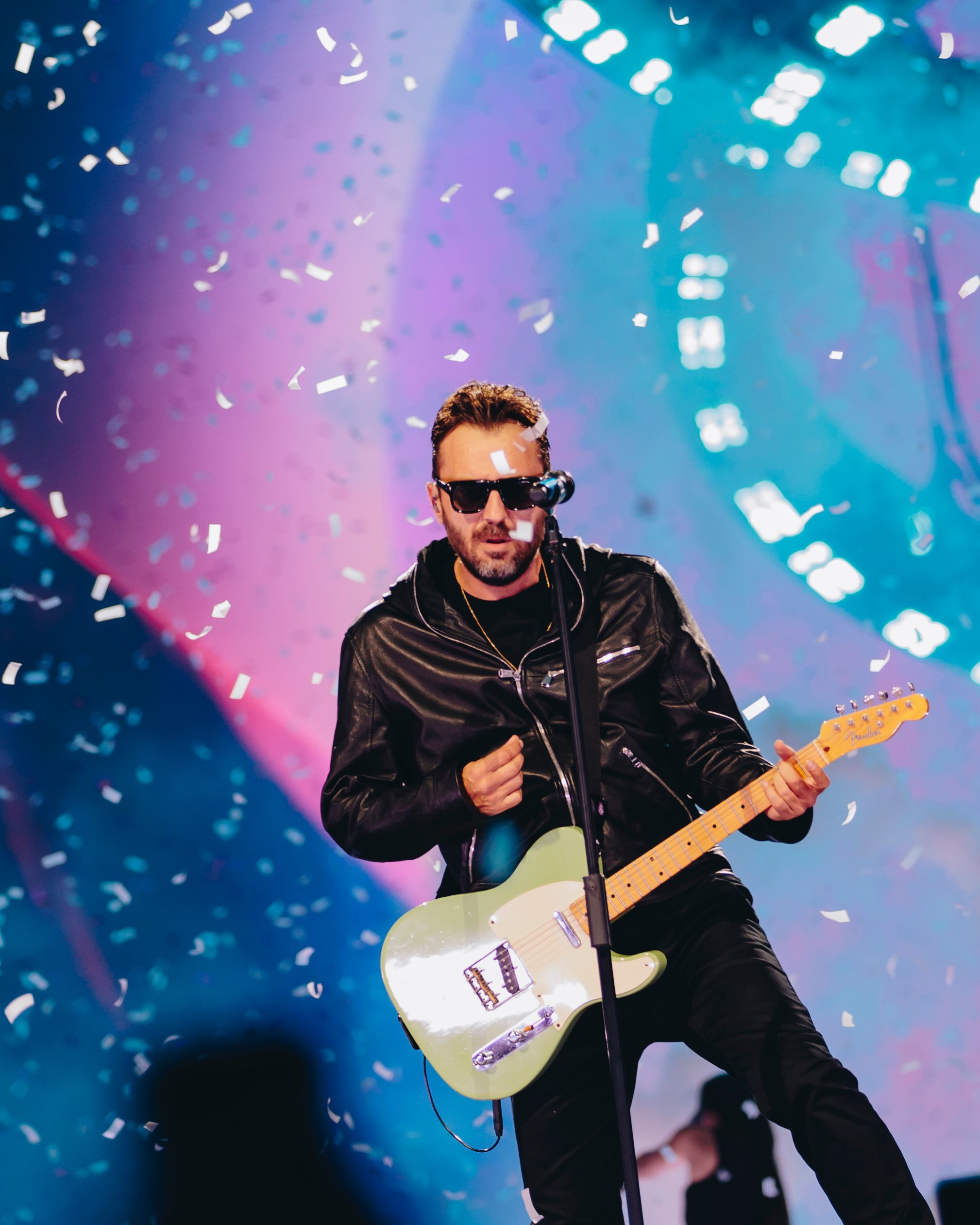 Cesare Cremonini in concerto, vestito di nero e con gli occhiali da sole. Sta suonando una chitarra elettrica, sullo sfondo ci sono degli schermi colorati di blu e viola e davanti volano dei coriandoli.
