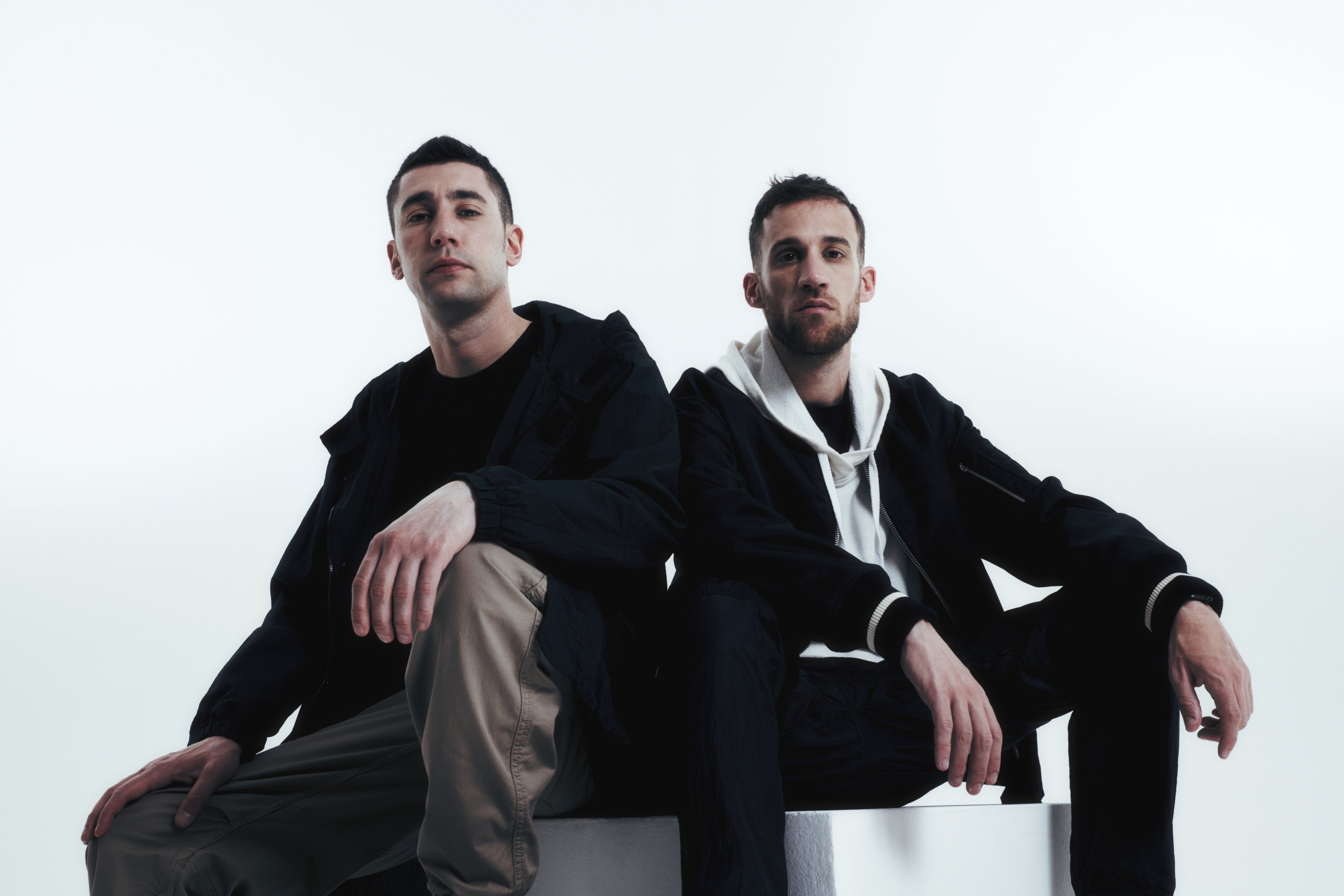 Marz e Zef seduti spalla a spalla su un piedistallo bianco, su sfondo bianco.