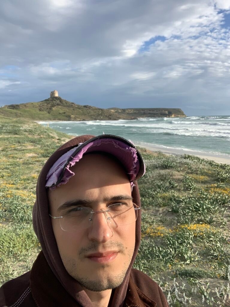 Selfie di Generic Animal con una felpa marrone col cappuccio tirato sul un cappellino con la visiera un po' sgualcito. Sullo sfondo una costa, il mare, ed una torre in lontananza.