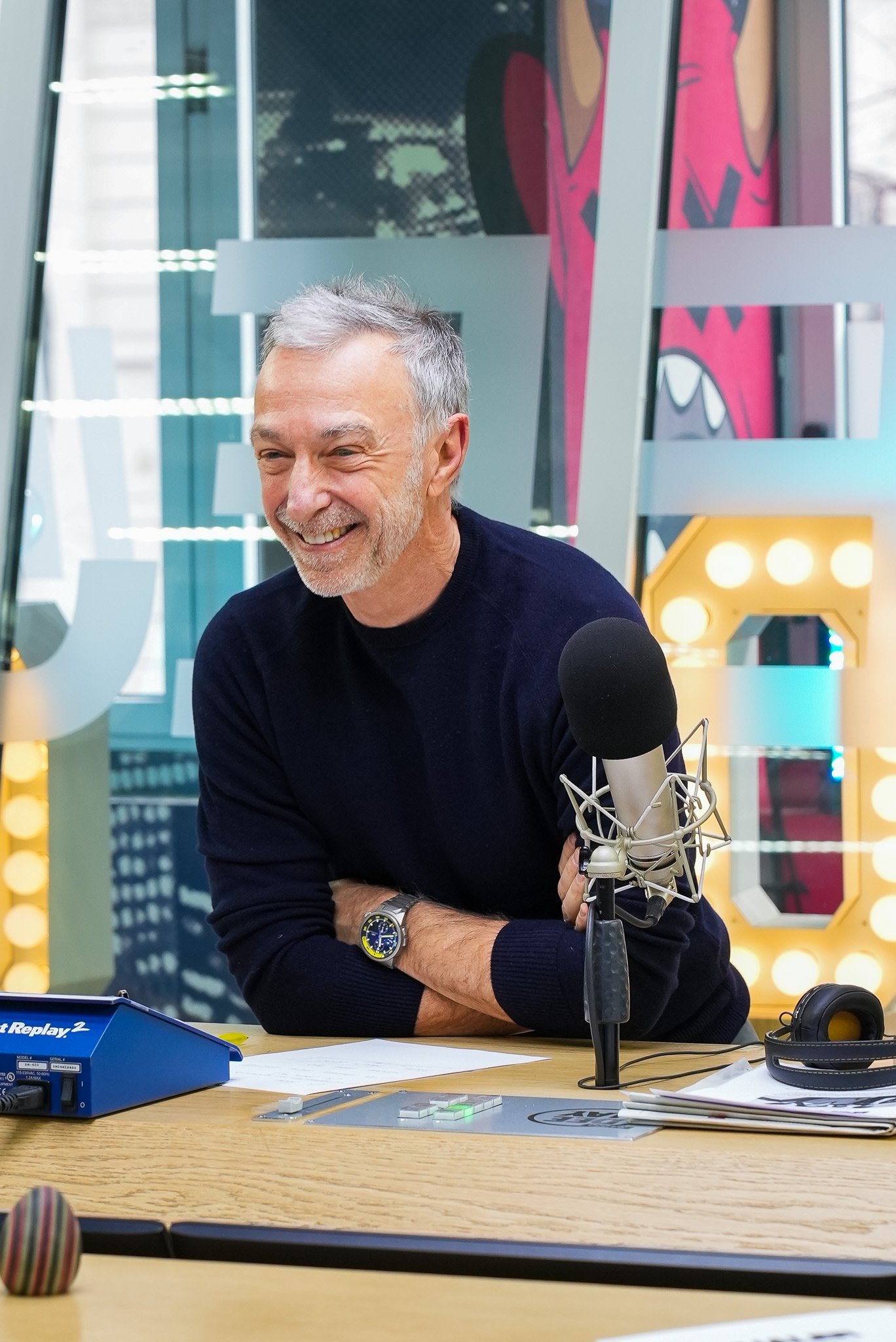 Linus che sorride appoggiato al tavolo dello studio di registrazione, con un microfono vicino.